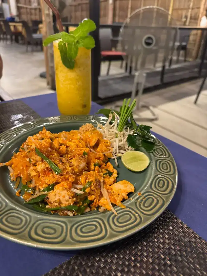 Pad thai with lime and bean sprouts on a green plate, and a tall yellow drink with mint