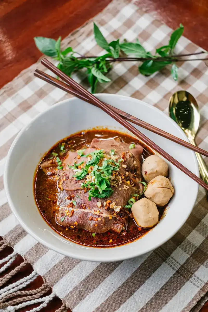 Boat noodles with sliced beef, three beef balls and chopped herbs, a sprig of Thai basil and a gold spoon on a brown checked cloth