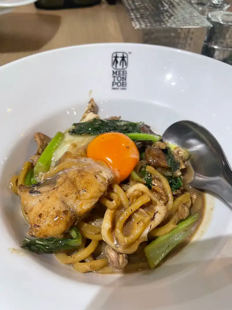 Fried yellow noodles with seafood, greens and an egg yolk, in a white bowl stamped Mee Ton Poe since 1946