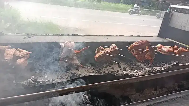Chickens on skewers in a row over a long charcoal trough, with the road behind
