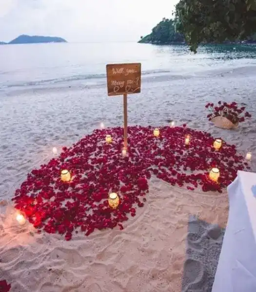 Rose petals and candles laid out on the sand around a small wooden sign, the sea behind