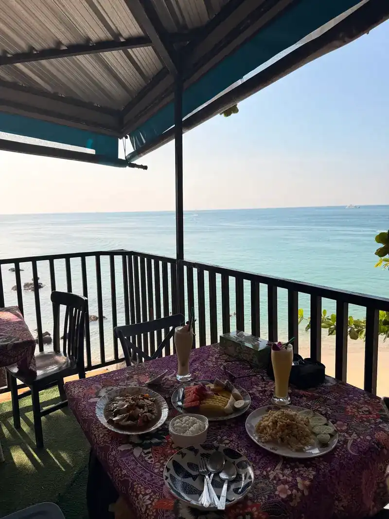 A table set with several dishes and drinks on the covered deck, the sea behind the railing.