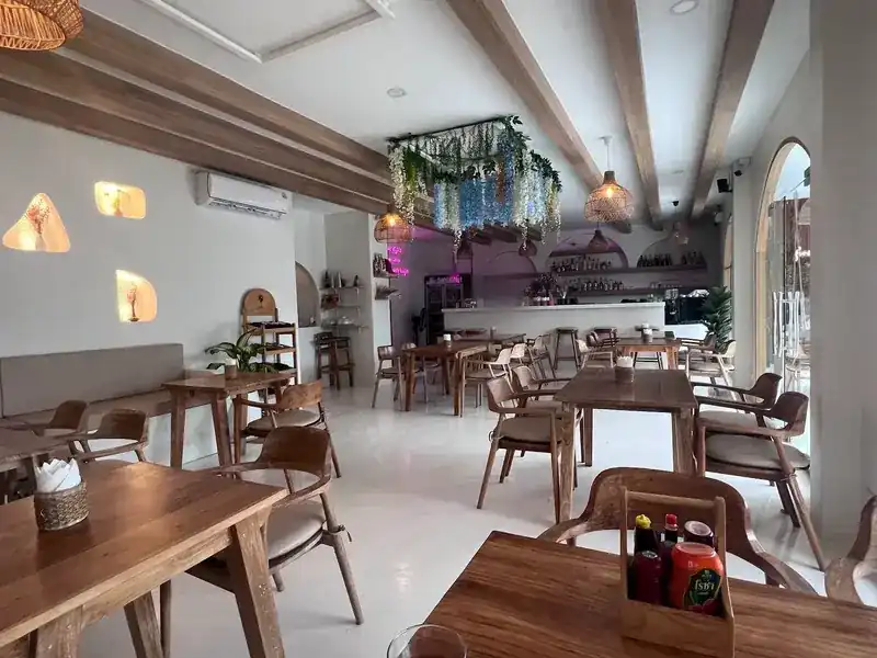 The dining room: wooden tables and chairs, white walls with lit niches and the bar at the back