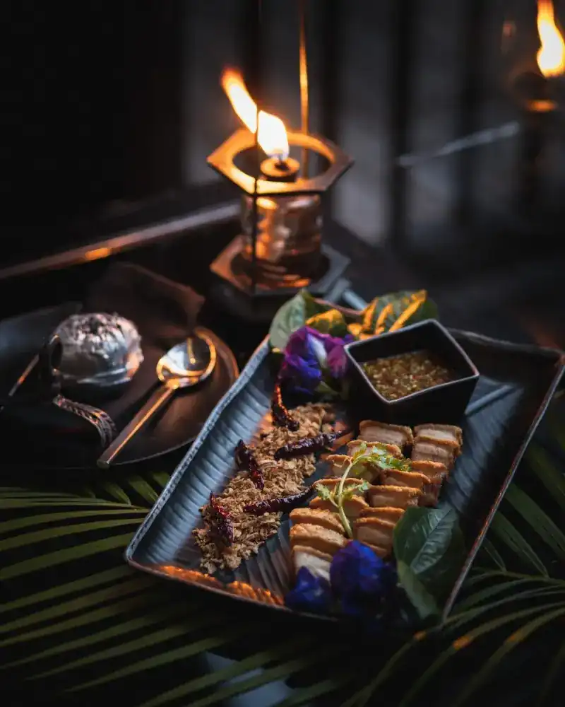 Crisp slices of pork with a chilli dip and blue butterfly-pea flowers on a long dark platter