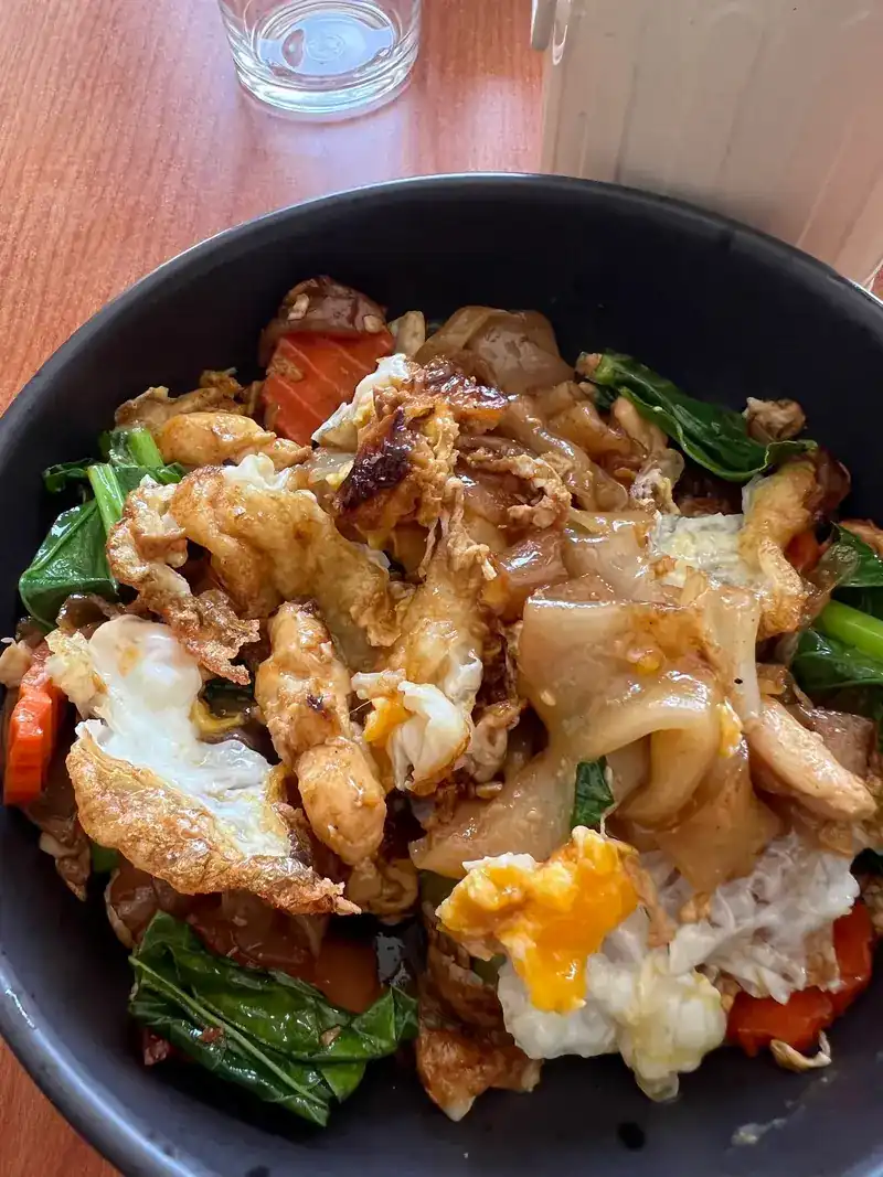 Wide rice noodles stir-fried with chicken, greens, carrot and a fried egg in a dark bowl