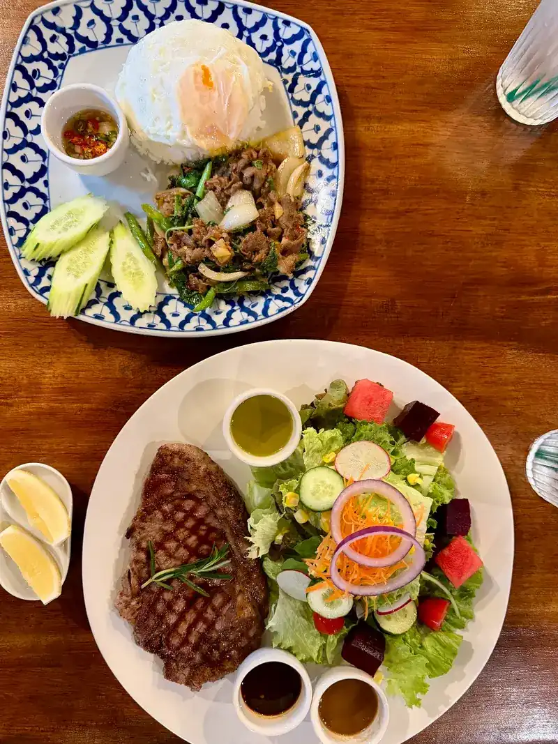 Seen from above: a steak and a large salad on one white plate, with stir-fried beef on rice and a fried egg on a blue and white plate behind