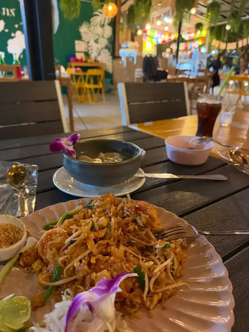 Pad thai with prawns, lime and crushed peanuts, with a bowl of dumplings in broth behind it.