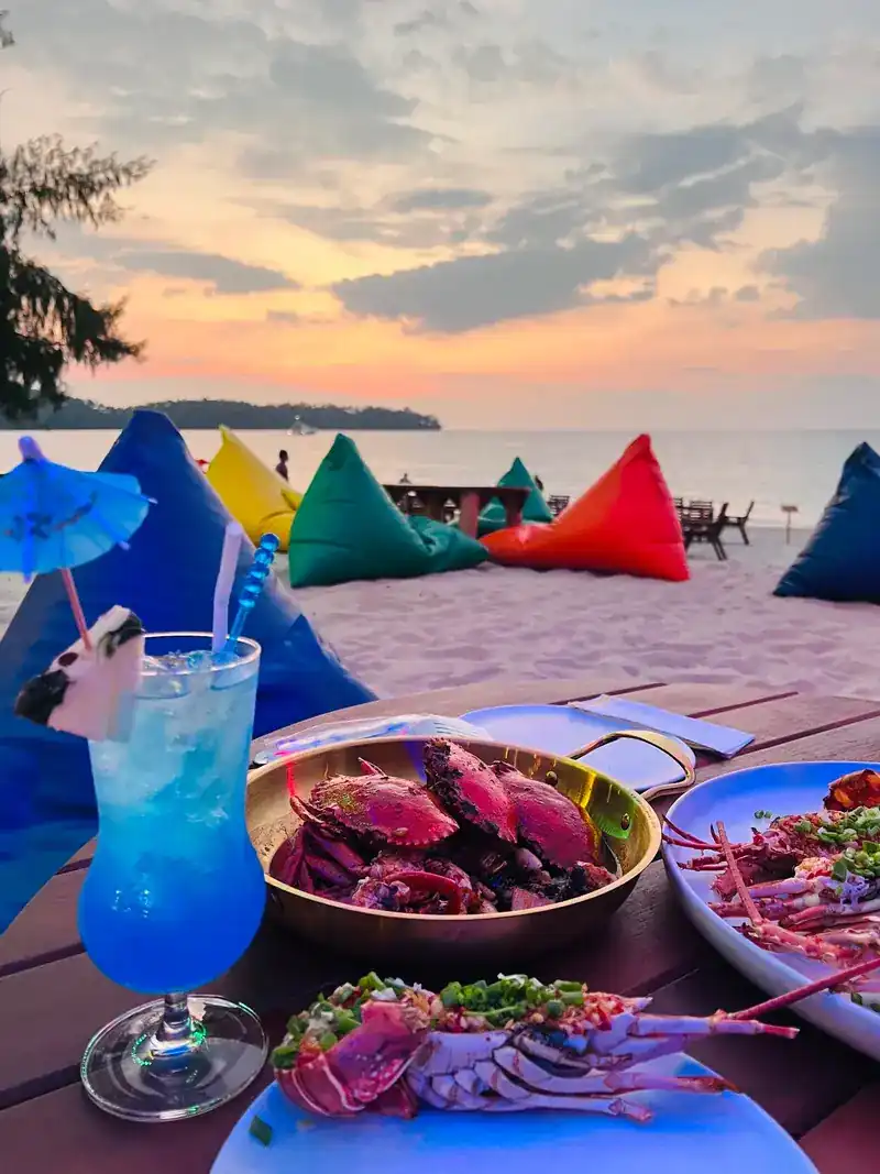 Crabs, lobster and a blue drink on a table, colourful beanbags on the sand and the sea at sunset