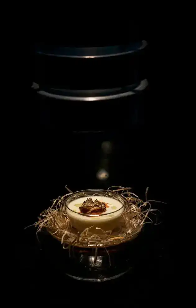A small glass bowl of cream topped with truffle, set in a nest of straw, lit in the dark