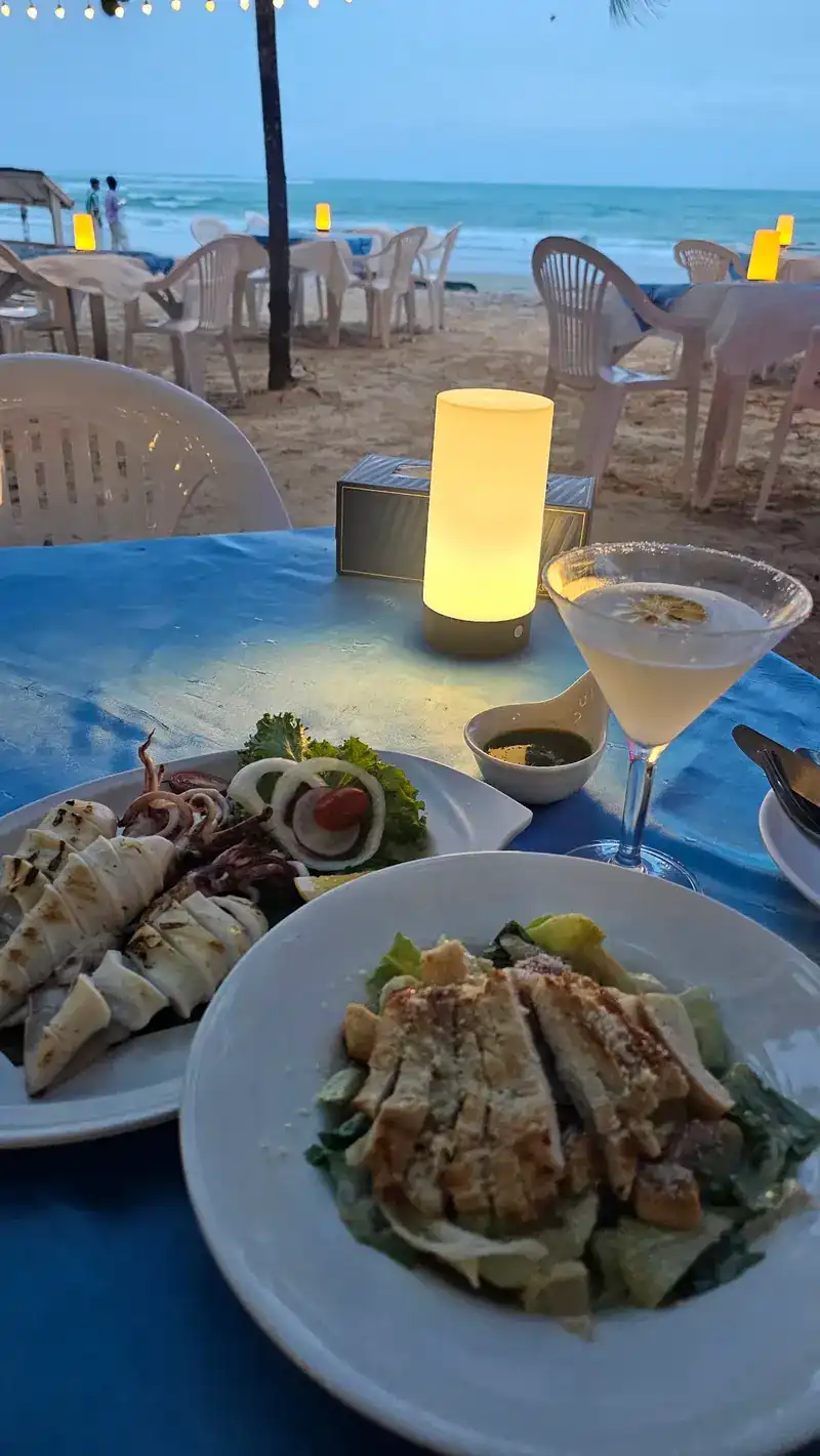 A table on the sand at dusk with a lit lamp, a plate of squid, a salad and a glass, the sea behind