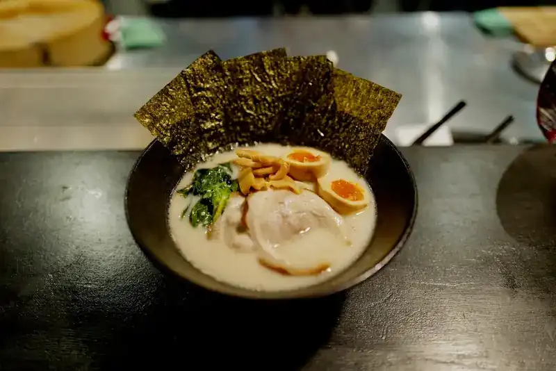 A bowl of ramen in creamy broth with soft eggs, greens and sheets of nori, on a dark counter / ราเมงน้ำซุปข้นกับไข่ยางมะตูม ผักและสาหร่ายแผ่น บนเคาน์เตอร์สีเข้ม