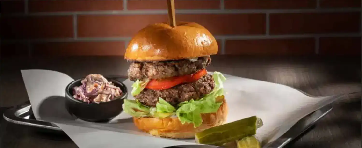 The Godfather: a double beef patty burger with tomato and lettuce, coleslaw and a pickle, on a tray against a brick wall