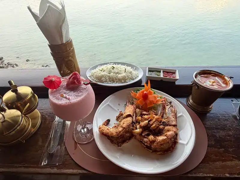 Two big tiger prawns on a white plate with rice, a curry in a brass pot and a pink drink, the sea below the railing.