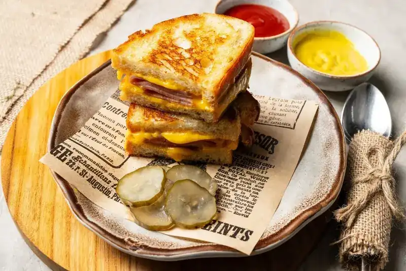 A toasted ham and cheese sandwich stacked on printed paper, with pickles, ketchup and mustard