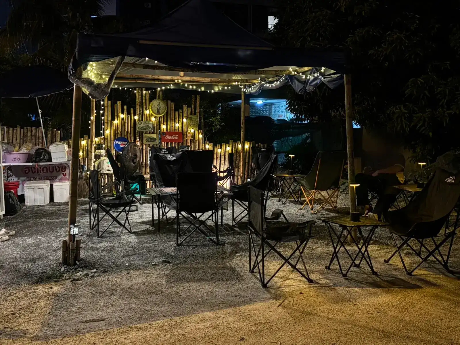The shop at night: camp chairs and small tables on gravel under a canopy strung with fairy lights, in front of a lit bamboo wall