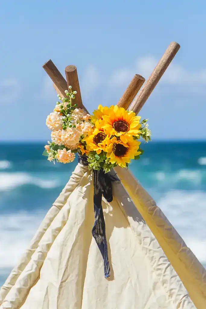 A canvas tipi tied with sunflowers on the beach, waves behind