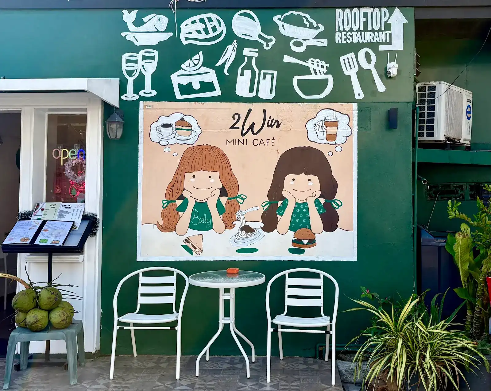 The green front of 2Win Mini Café: a painted wall with two girls, white chairs outside and a sign pointing up to the rooftop restaurant