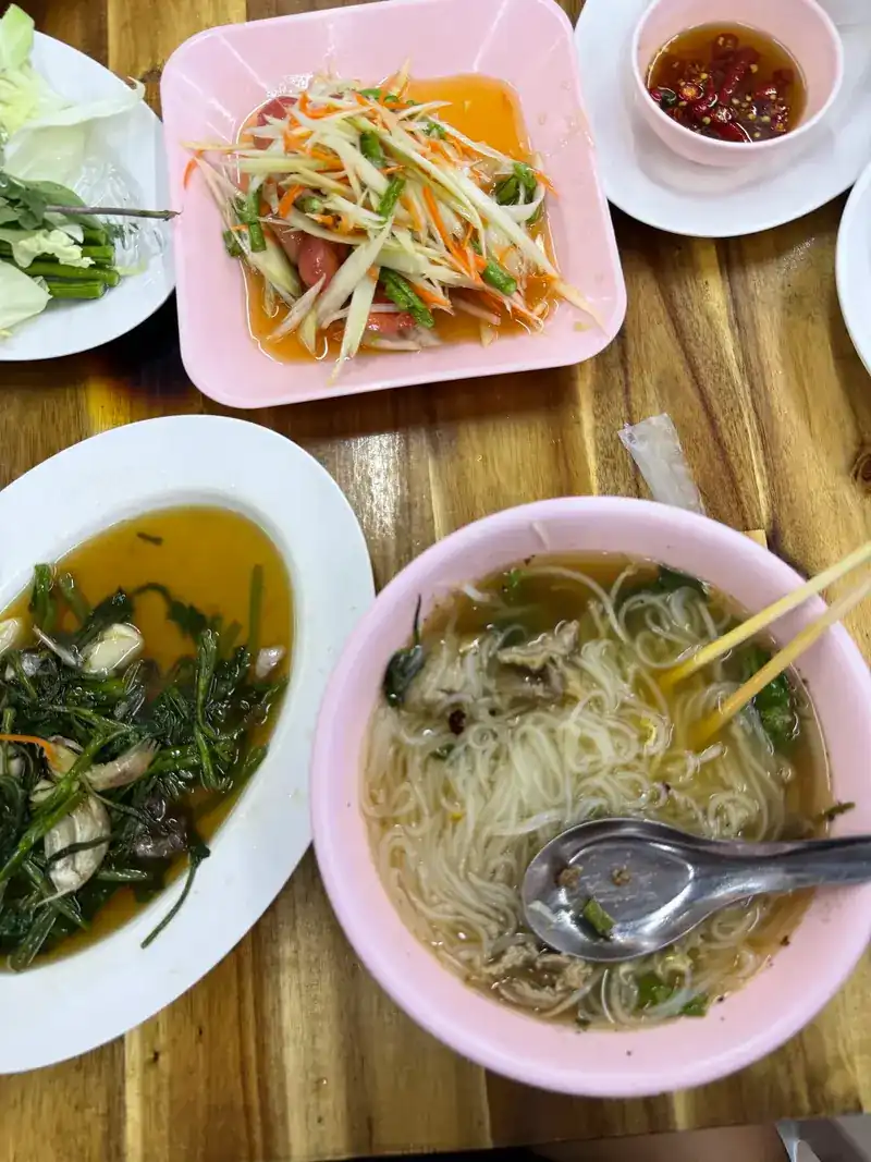 A spicy shredded salad, a bowl of greens and a bowl of noodle soup on a wooden table