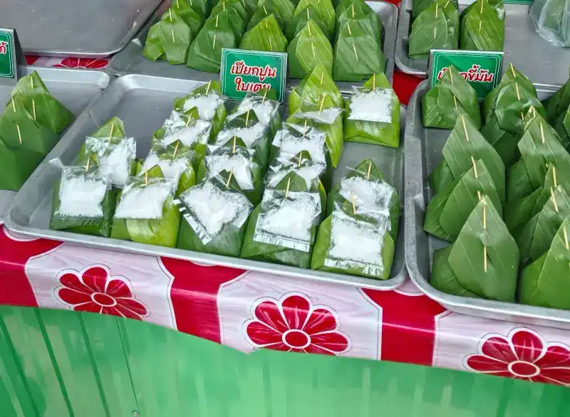 Banana-leaf parcels pinned with bamboo on steel trays, over a red flowered tablecloth