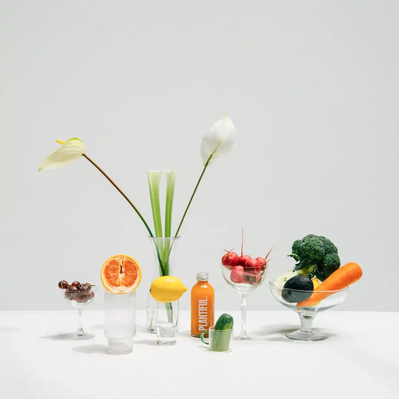 A still life on a white table: grapefruit, lemon, radishes, broccoli, a carrot and a bottle of Plantiful juice