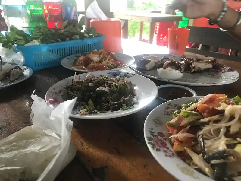 A wooden table with papaya salad, minced-meat salad, grilled chicken, sticky rice and a basket of fresh greens