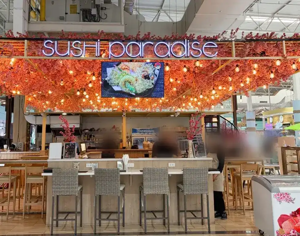 The Sushi Paradise counter: a neon sign in a canopy of coral blossom and warm bulbs, above a row of high stools