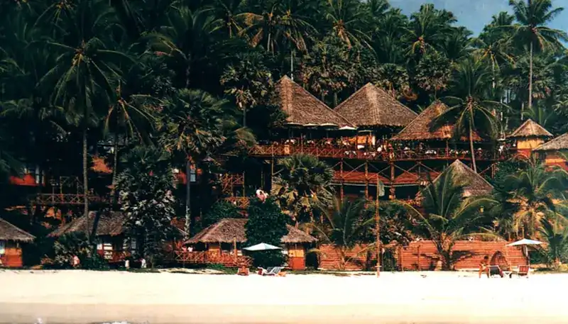 An old photograph of thatched pavilions on wooden decks among palms above the beach, from the hotel's early years as The Pansea