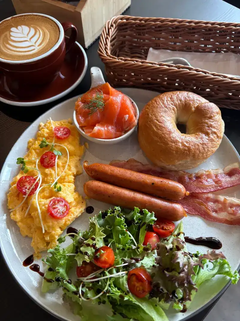 A breakfast plate with a bagel, smoked salmon, scrambled eggs, sausages, bacon and salad, next to a latte
