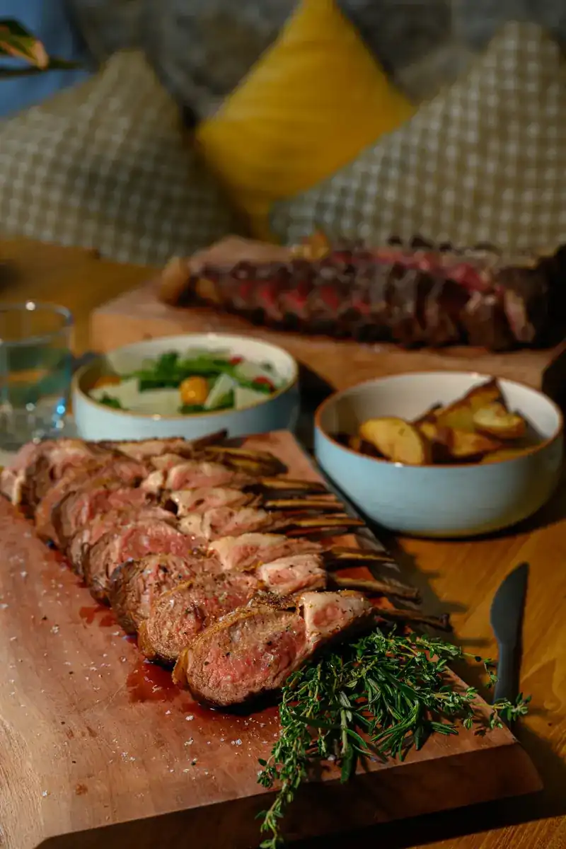 Sliced rack of lamb on a wooden board with thyme, roast potatoes and salad