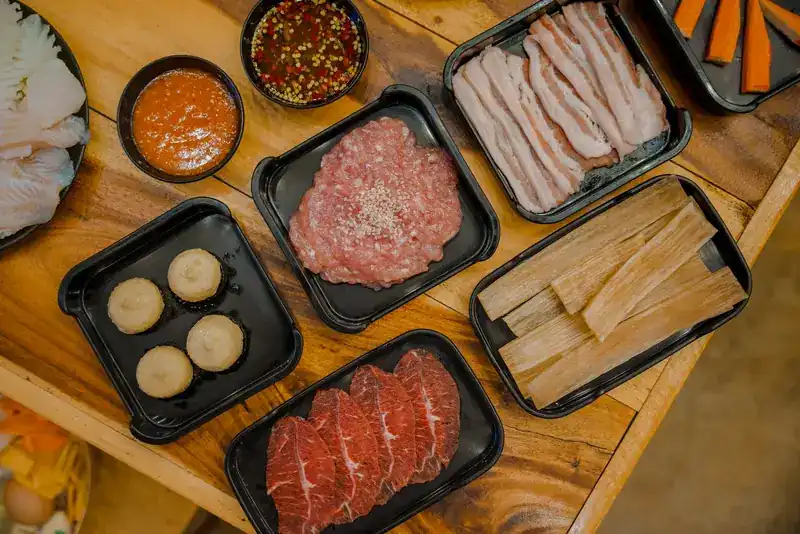 Black trays of pork belly, minced pork, beef and mushrooms with two dipping sauces, seen from above