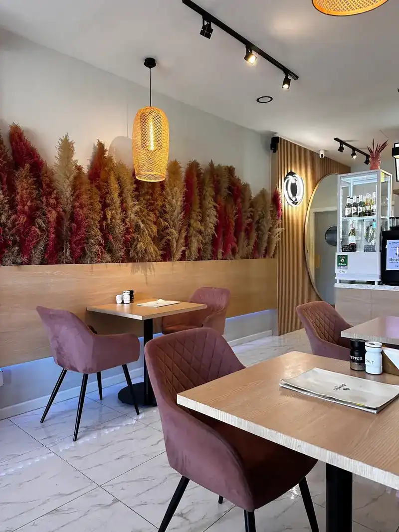 Inside the cafe: a wall of red and gold pampas grass above wooden tables and dusty-pink chairs, under a rattan lamp