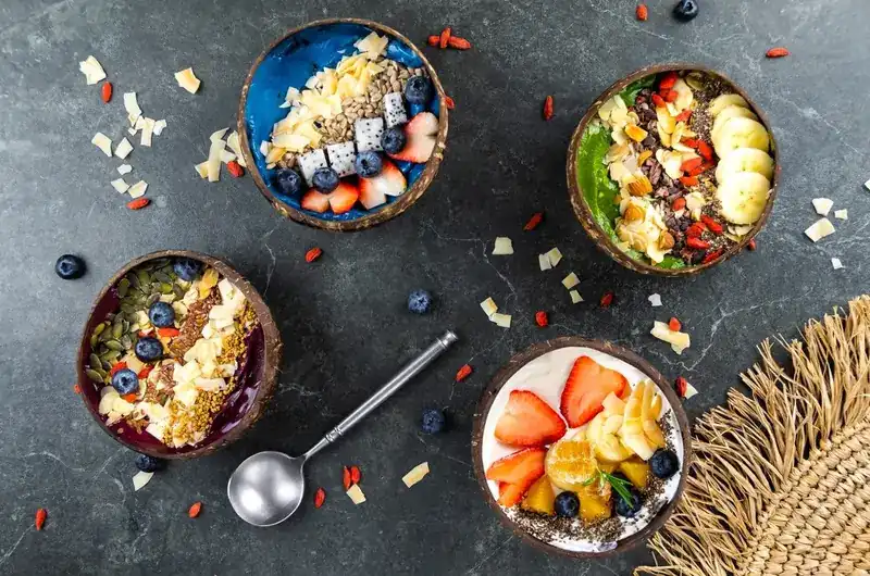 Four smoothie bowls in coconut shells topped with fruit, seeds and coconut flakes