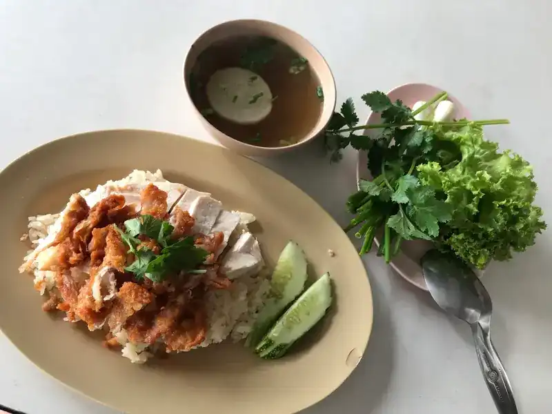 A plate of boiled and fried chicken over rice, with clear soup, cucumber, lettuce and coriander
