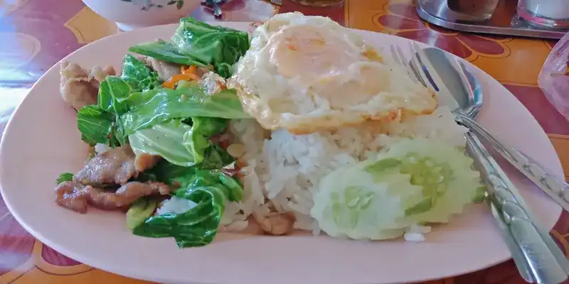 Stir-fried greens with pork, rice, a fried egg and sliced cucumber on an oval plate