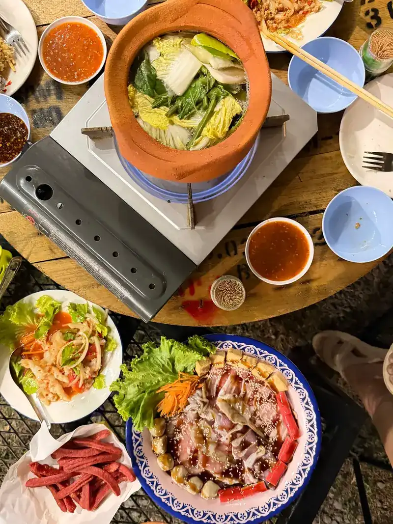 Jim jum seen from above: a clay pot of broth and vegetables on a gas burner, with papaya salad, dipping sauces and a plate of sliced meat