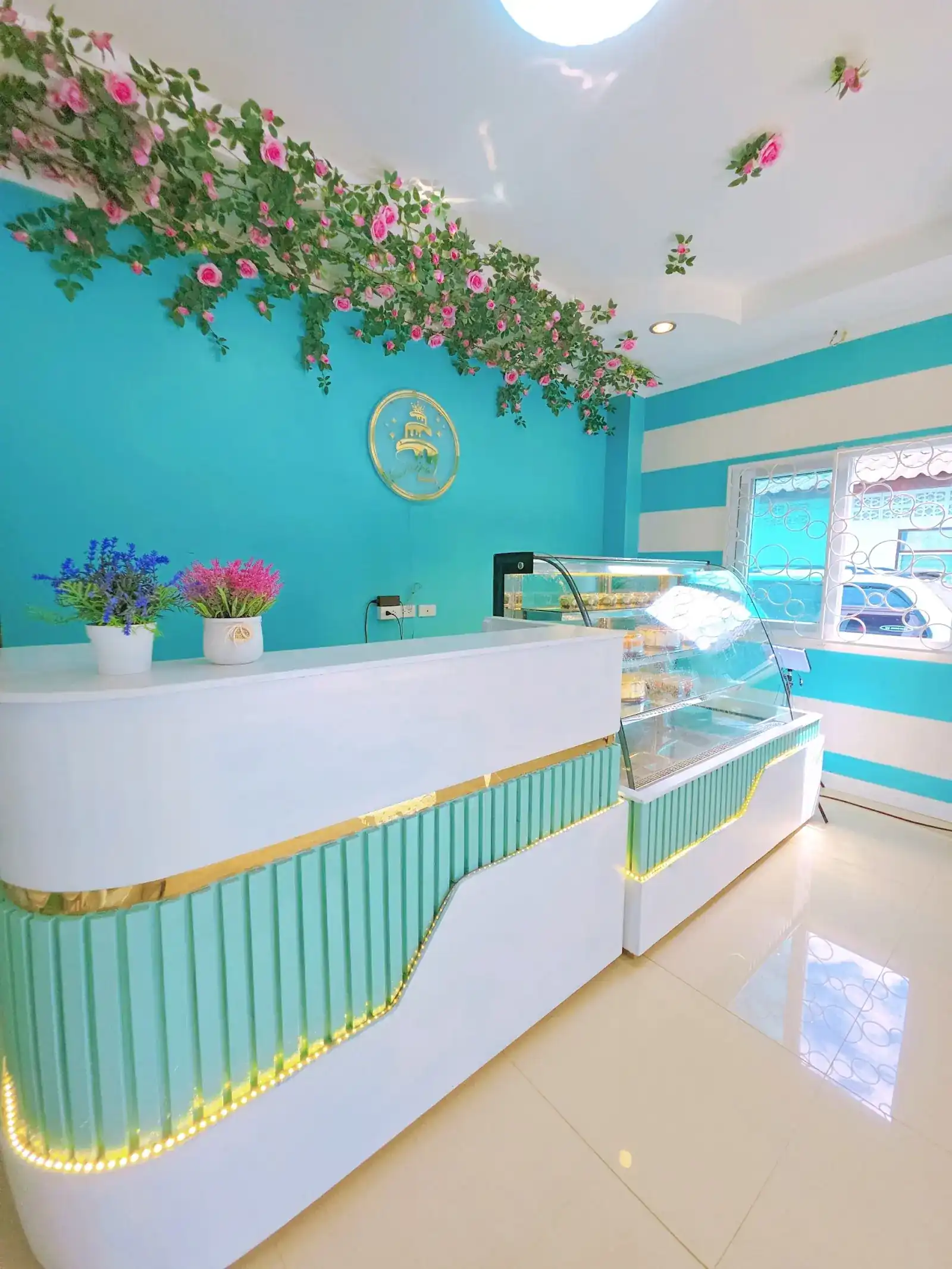 The white counter with its mint fluted front and gold trim, a glass cake case, and pink roses along the turquoise wall