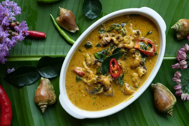 A coconut curry in a white dish on a banana leaf, with shells, chillies and flowers around it
