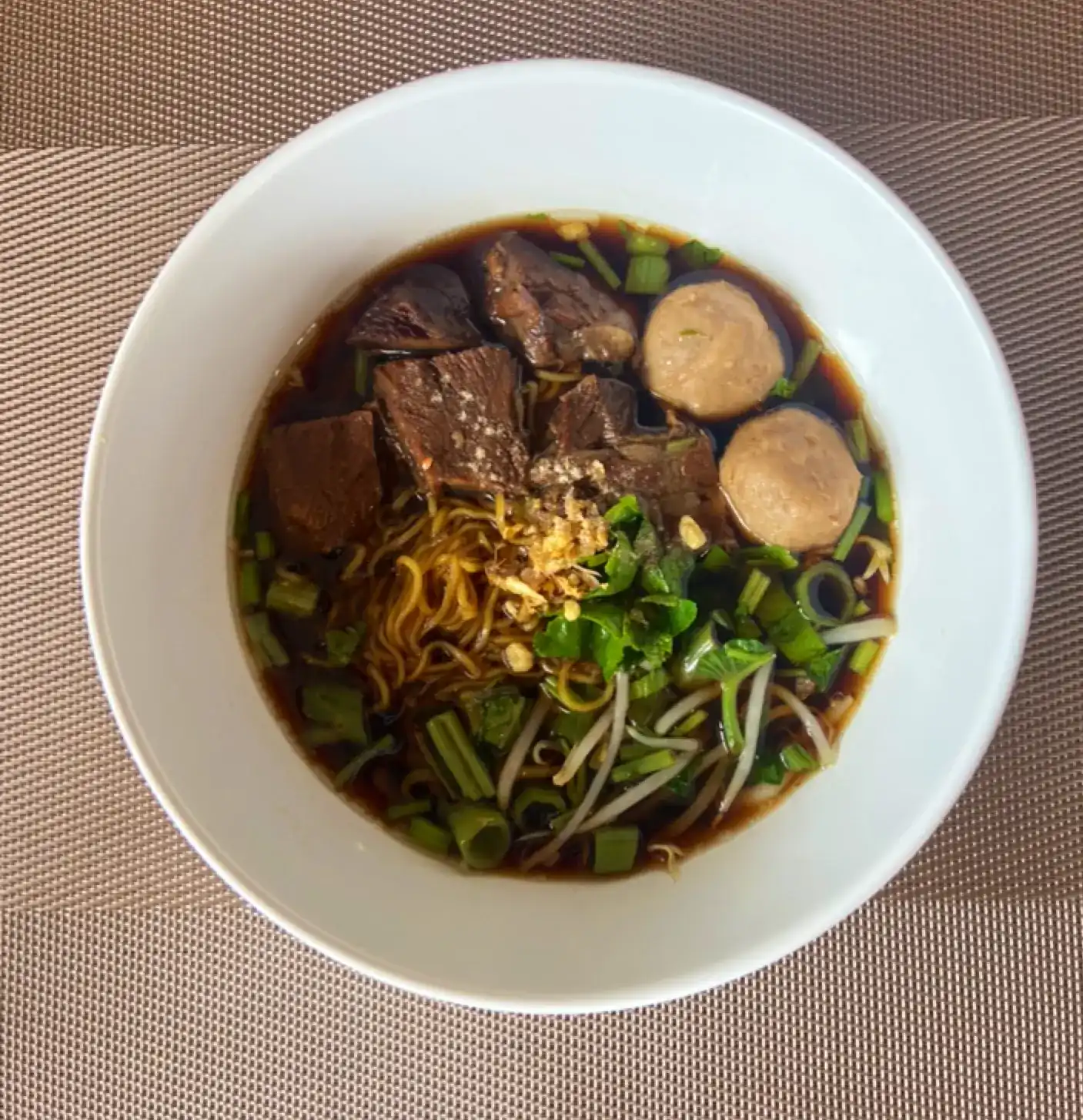 A bowl of egg noodle soup with braised beef, meatballs, greens and bean sprouts