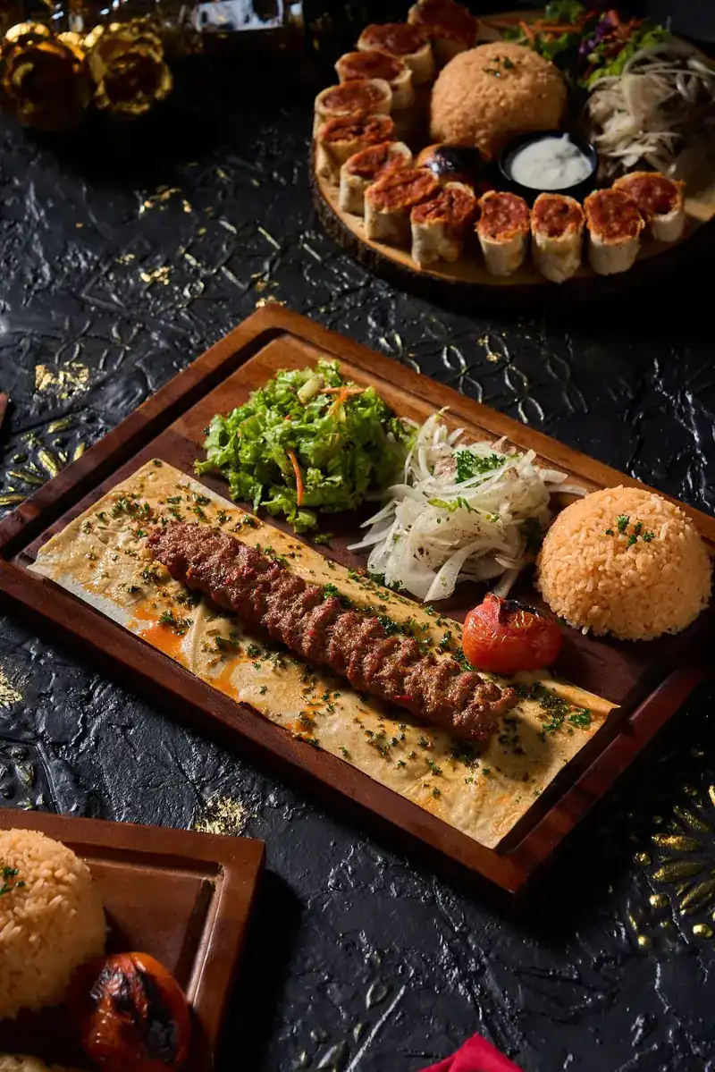 A long grilled kebab on flatbread with salad, onions, rice and a charred tomato
