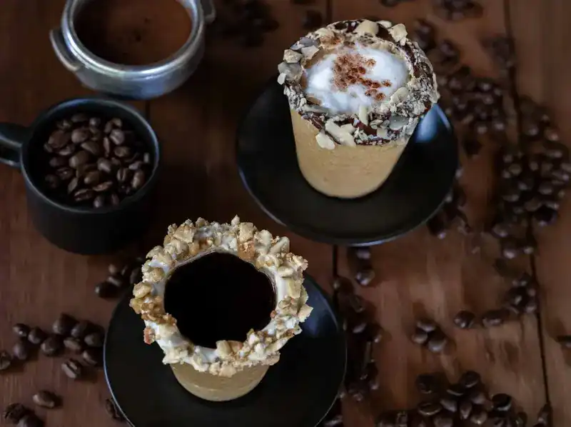 Two edible cups with nut-crusted rims, one with black coffee and one with milk foam