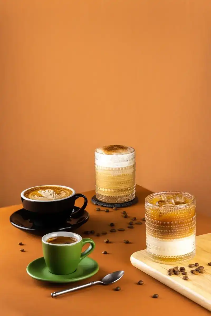 A latte in a black cup and two iced coffees against an orange wall