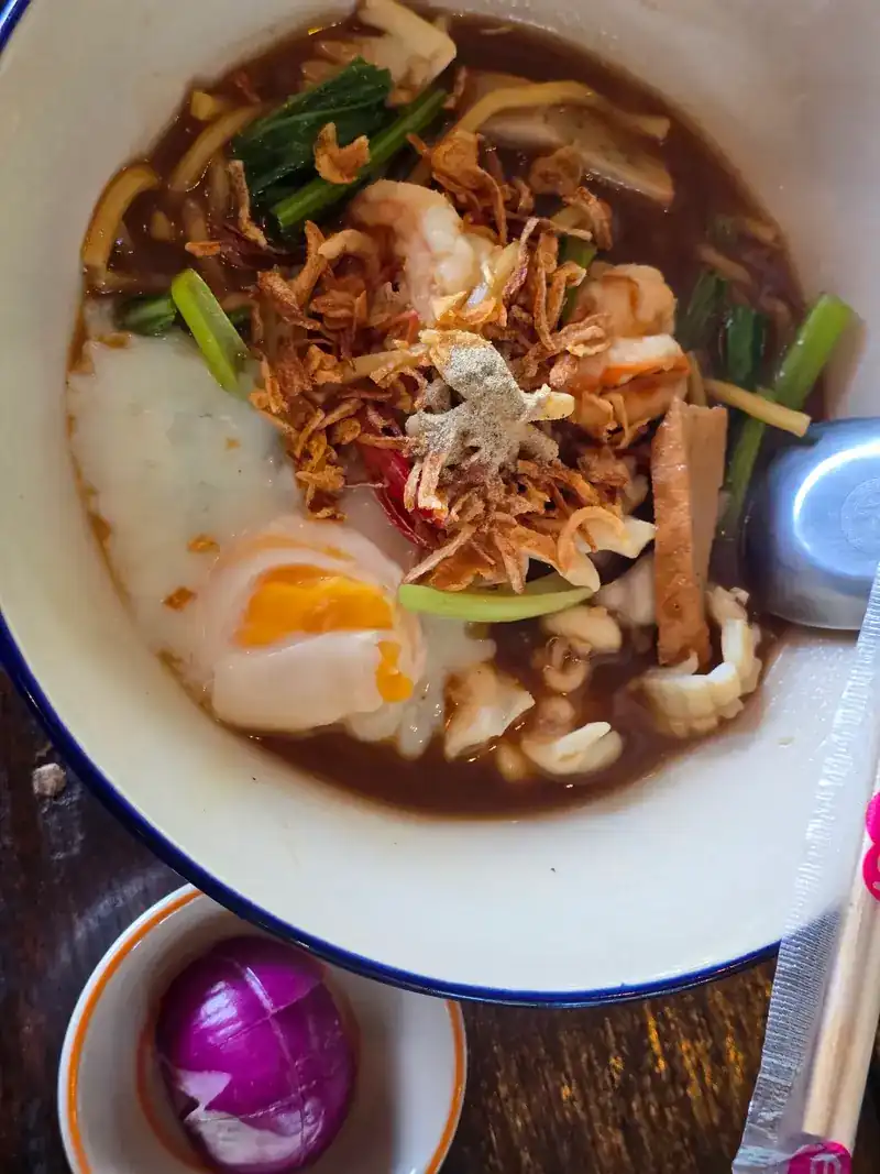 A bowl of yellow noodles in broth with seafood, a soft egg and fried shallots