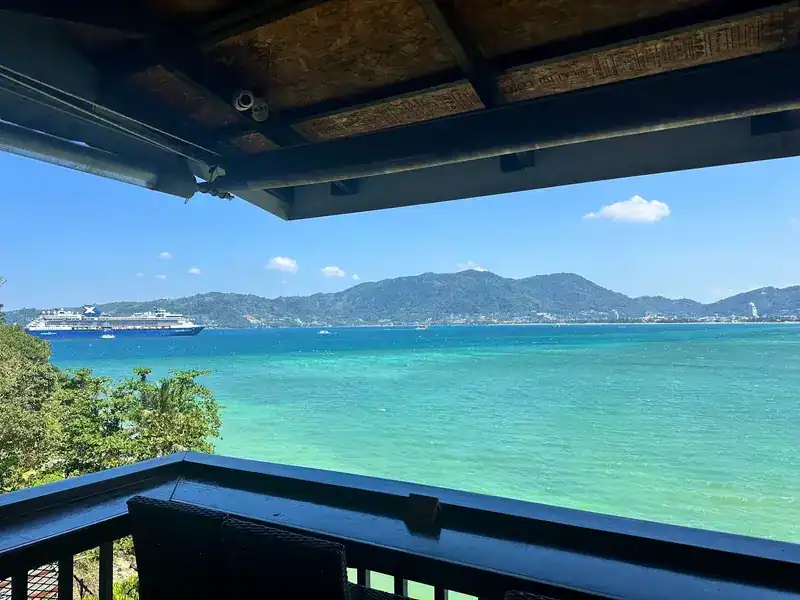 From under the terrace roof: turquoise sea, a cruise ship and green hills across the bay.