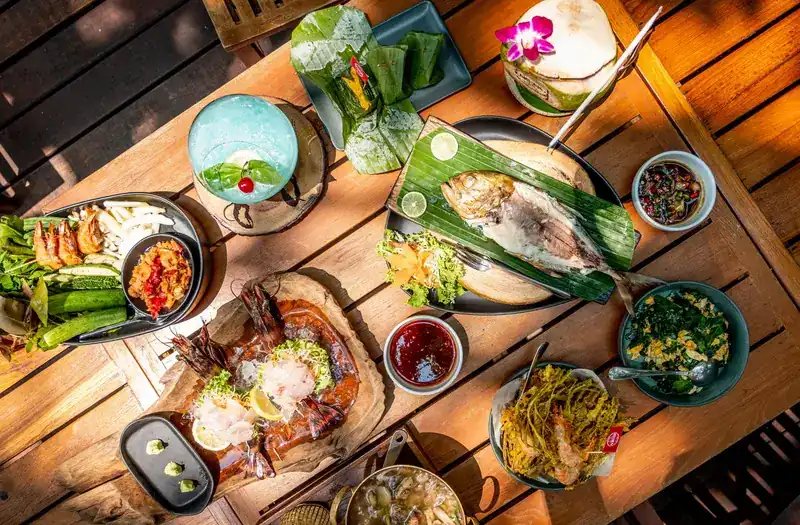 A wooden table seen from above with grilled fish on a banana leaf, prawn sashimi, chili dip with vegetables and fried freshwater weed