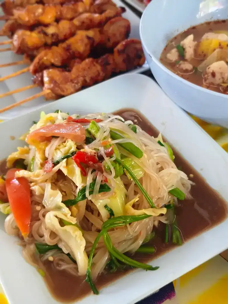 Papaya salad with noodles, with chicken skewers and a bowl of soup behind