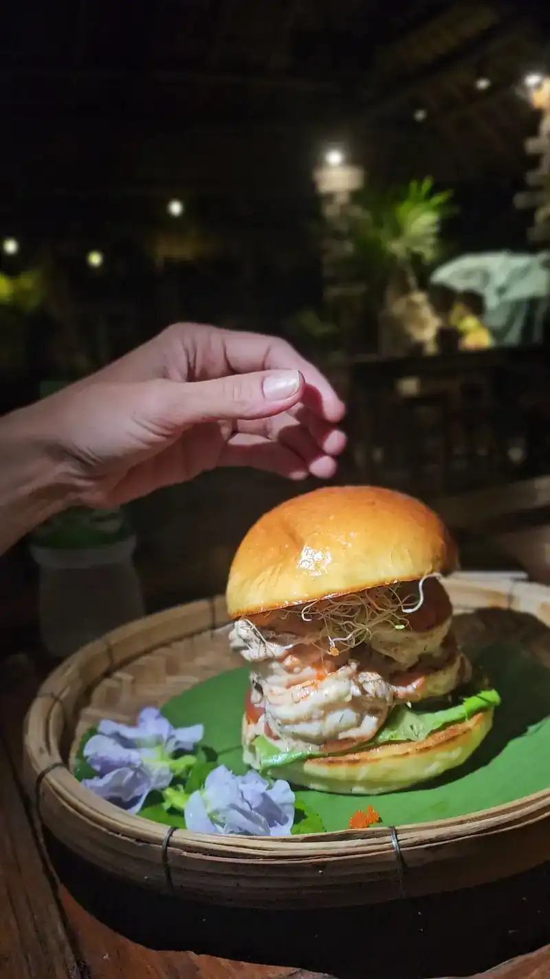 A burger in a bamboo tray at night under the garden lights, a hand reaching for it