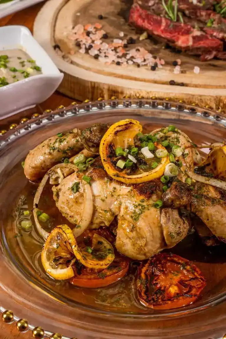 Roast chicken with charred lemon and tomatoes in a glass dish, steak on a wooden board behind