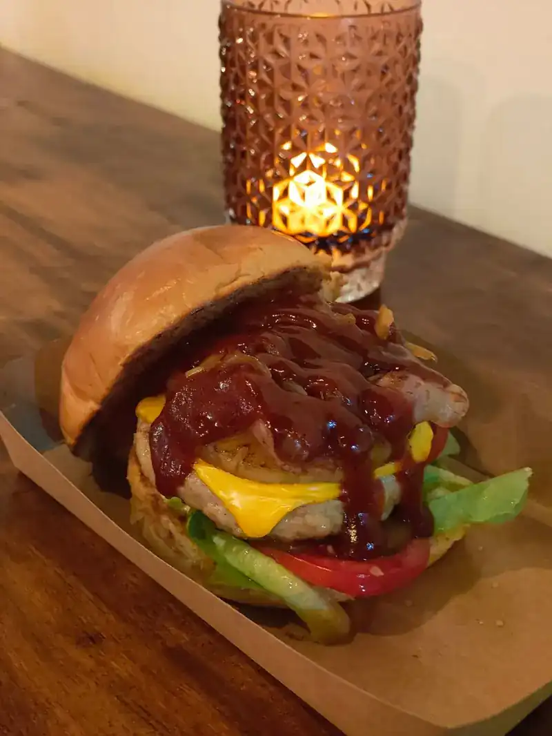 Burger with barbecue sauce, cheese, tomato and lettuce in a paper tray, a candle glowing behind it
