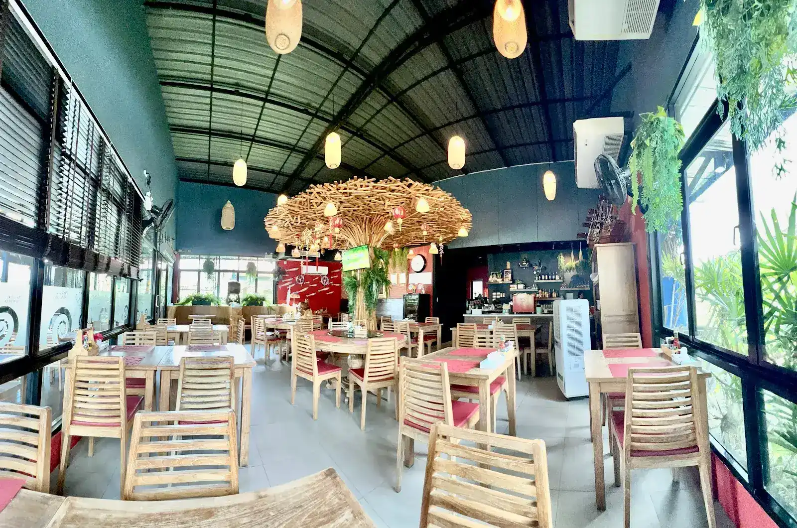 The dining room: wooden chairs with red cushions under a large canopy of wooden sticks and hanging lamps, glass walls and plants