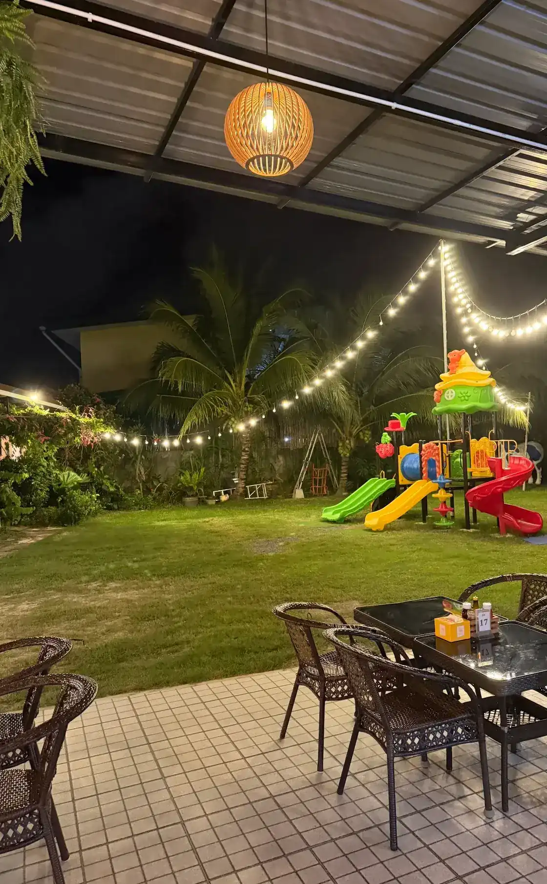 Terrace tables and rattan chairs under a hanging lamp, looking out at night over a lit lawn with a children's slide set and palm trees.